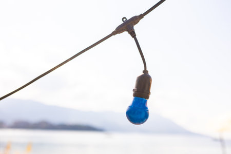 Blue light bulb hanging above beach sand. Decorative lighting element with blurred seaside background and wooden terrace.の写真素材