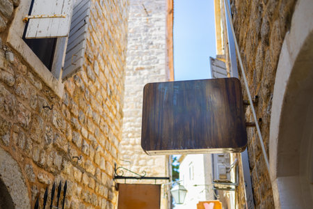 Blank wooden sign hanging between stone walls in narrow Mediterranean alley, copy space. Street branding, tourism, and vintage commercial aesthetics in coastal old towns.の写真素材