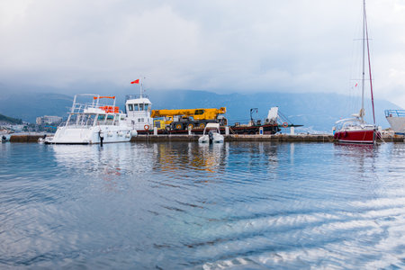 Boats docked at harbor under cloudy sky with construction vehicle on pier. Marina transport, nautical lifestyle and industrial elements by the sea.の写真素材