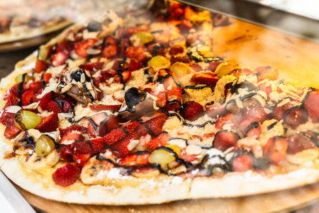 Colorful fruit dessert pizza behind glass. Sweet toppings, gourmet snack, and indulgent street food with fresh strawberries, banana slices, and melted chocolate.の写真素材