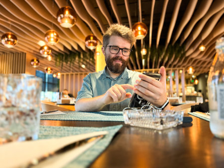 Man using smartphone at dining table in restaurant. Lifestyle, communication, leisure and casual technology interaction within modern dining interior and social atmosphere.の写真素材