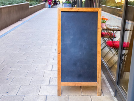 Wooden chalkboard sign standing on a pedestrian street next to cafe terrace and pink chairs. Urban commerce, communication and business display symbolizing customer service and advertising copy space.の写真素材