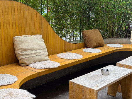 Wooden curved bench with pillows and small table placed in outdoor terrace. Hospitality, leisure, and lifestyle expressed through decor, design, and natural greeneryの写真素材
