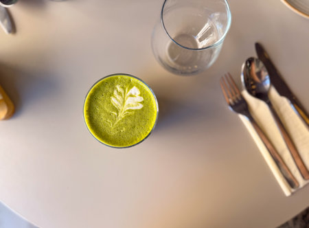 Glass of matcha latte with leaf foam art served on cafe table in soft daylight top view. Healthy...