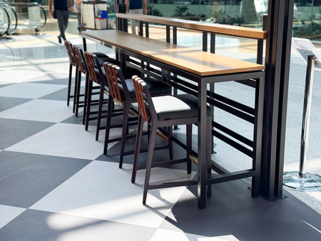 Tall wooden tables with high stools arranged by large windows in a public lounge. Representation of travel lifestyle, dining, and social atmosphere in modern hospitality spaces.の写真素材