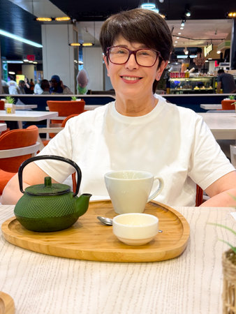 Woman sitting at cafe table with teapot and cup smiling warmly. Social lifestyle, hospitality, and leisure highlighting dining moment, communication, and cheerful mood.の写真素材