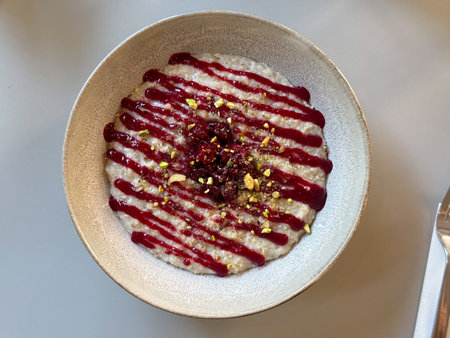 Bowl of creamy oat pudding with raspberry sauce and crushed pistachios served on table. Dessert,...
