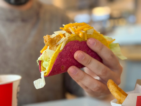A person holds a colorful taco with cheese, lettuce and crispy filling inside a cafe. Fast food culture, casual dining, consumer lifestyle and modern gastronomy in urban environment.の写真素材