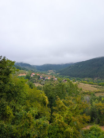Foggy mountains and a valley with small houses and red roofs among green trees. Peaceful rural...