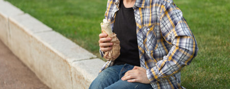 Man holding a street food wrap in craft paper outdoors. Concept of fast food, casual lifestyle, and outdoor snacking in urban park settings. Banner with copy spaceの写真素材