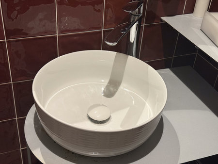 Close-up of round white sink and chrome faucet against glossy red tiled wall. Modern bathroom...