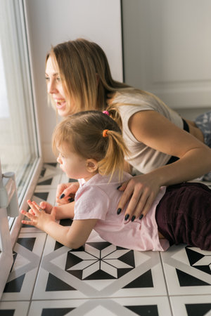 Mother and daughter lying together by the window and watching the outside world. Calm connection, curiosity, and the beauty of simple family moments at home.の写真素材
