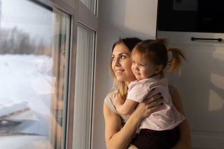 Mother holding her daughter close while watching through the window in soft sunlight. Family love, emotional peace, and gentle connection in quiet home atmosphere.の写真素材
