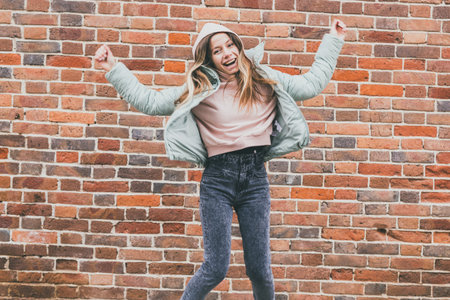 Smiling funny woman in mint jacket jumping joyfully near brick wall. Spontaneity, happiness, and...