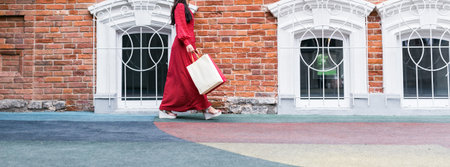 Woman in red polka dot dress walking with shopping bag near vintage brick building Banner with copy space and empty place for advertising. Fashion, city lifestyle, and elegance in urban environment.の写真素材