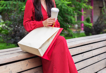 Woman in red polka dot dress sitting with coffee cup and shopping bags outdoors. Relaxation, consumer lifestyle, and leisure concept.の写真素材