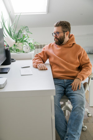 Man sitting at desk in bright home office. Scene symbolizing focus, reflection, technology use and professional routine in contemporary domestic workspace.の写真素材