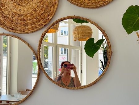 Woman taking mirror selfie in bright cafe interior. Natural light, reflection, and candid everyday lifestyle moment.の写真素材