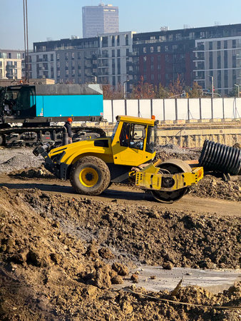 Construction roller moving across soil mound near residential buildings. Infrastructure development,...