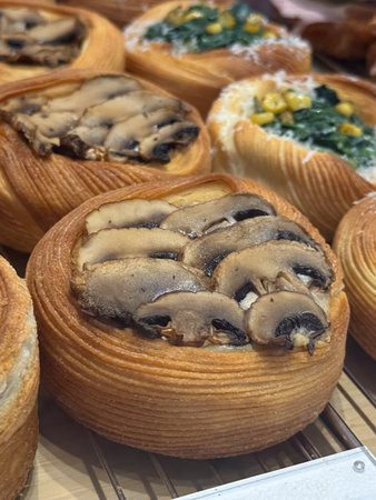 Freshly baked pastries with mushrooms and vegetables in bakery display close-up. Savory flavors,...