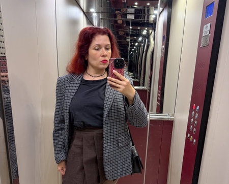 Woman in checked blazer and short brown skirt taking a selfie in elevator close-up. Expression,...