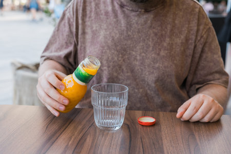 Person holding bottle of orange juice above glass at cafe table outdoors. Beverage preparation,...