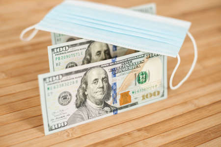 A medical blue mask lies on dollars with a face value of 100 dollars on a wooden background. 100...