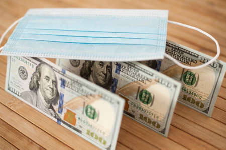 A medical blue mask lies on dollars with a face value of 100 dollars on a wooden background. 100 dollar bill and protection medical mask. Coronavirus. Financial assistance for medicine.の写真素材