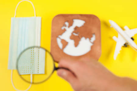 Plane model, face mask, loupe in hand and earth model on a yellow background. Flight impact of coronavirus (COVID-19) concept.の写真素材