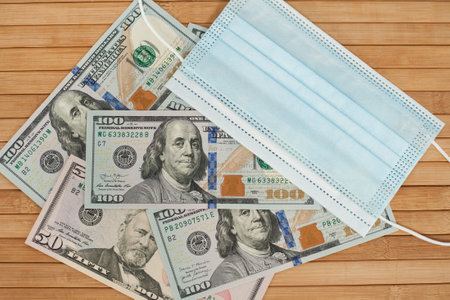 A medical blue mask lies on dollars with a face value of 100 dollars on a wooden background. 100 dollar bill and protection medical mask. Coronavirus. Financial assistance for medicine.の写真素材
