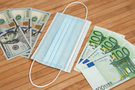 Money dollars, euro banknotes bill with face mask. Crisis and finance concept. COVID-19 coronavirus, pandemic. Mouth mask with euro and US dollar money on a wooden background.の写真素材