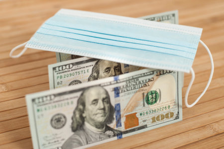 A medical blue mask lies on dollars with a face value of 100 dollars on a wooden background. 100 dollar bill and protection medical mask. Coronavirus. Financial assistance for medicine.の写真素材