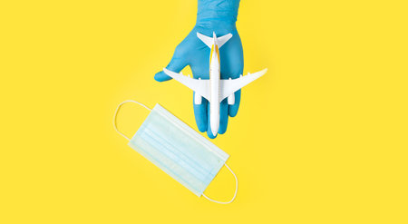 Hand in glove holding model airplanes on yellow background. Airplane model on hand and face medical mask Concept traveling. Copy spaceの写真素材