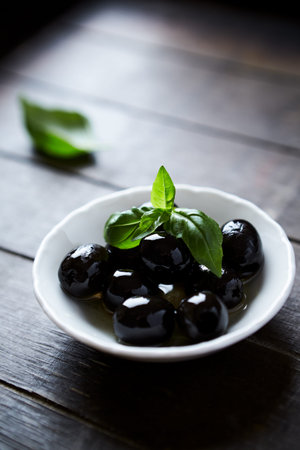 Black olives with basil leaves in a bowl on wooden table.