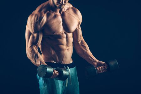 Closeup of a muscular young man lifting dumbbells weights on dark background .の写真素材