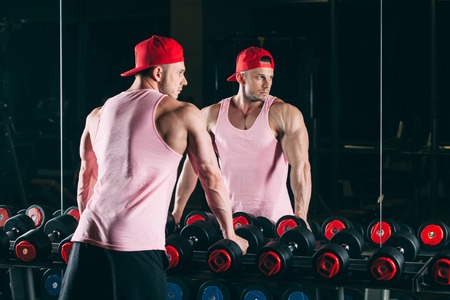 Muscular man out in gym standing near dumbbells, in a pink shirt and red baseball cap.の写真素材