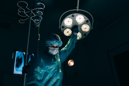 surgery, medicine and people concept - surgeon in mask adjusting lamp in operating room at hospitalの写真素材