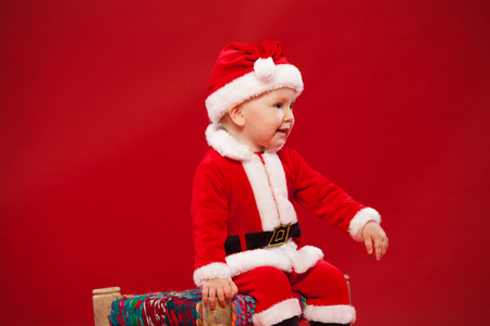 Beautiful little baby celebrates Christmas. New Year's holidays. Baby in a Christmas costume He is sitting on a chair on a red backgroundの写真素材