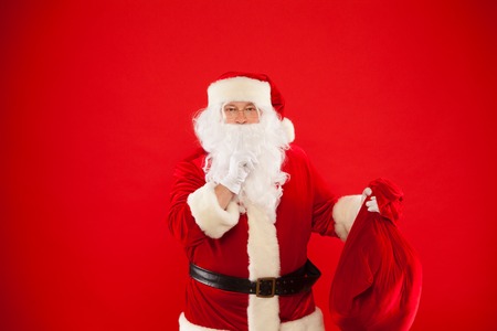 Portrait of Santa Claus with huge red sack keeping forefinger by his mouth and looking at camera. Merry Christmasの写真素材
