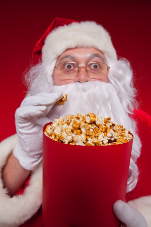 Close-up portrait Traditional Santa Claus watching TV, eating popcorn. Christmas. Red background. emotions. fear. surprise.の写真素材