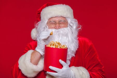 Traditional Santa Claus watching TV, eating popcorn. Christmas. Red background. emotions fear surpriseの写真素材