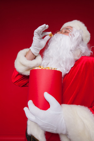 Close-up portrait Traditional Santa Claus watching TV, eating popcorn. Christmas. Red background. emotions. fear. surprise.の写真素材
