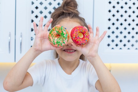 Close-up of pretty girl with double buns looking through the two sprinkled donuts on the background of bright kitchenの写真素材