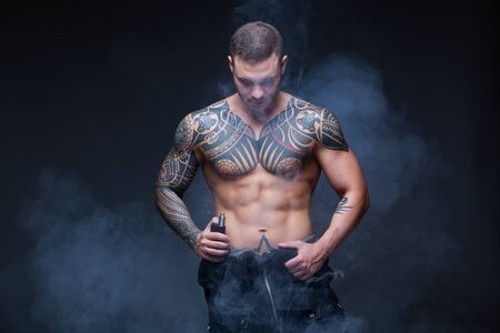 Vaper. The man with a muscular naked torso with tattoos smoke an electronic cigarette on the dark backgroundの写真素材