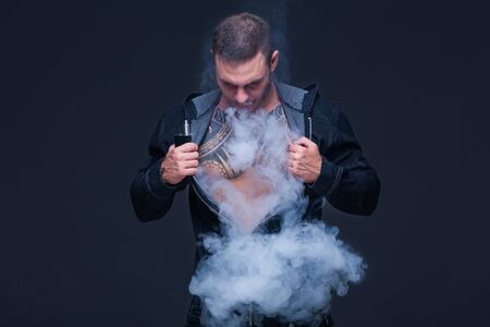 Vaper. The man with a muscular naked torso with tattoos smoke an electronic cigarette on the dark backgroundの写真素材