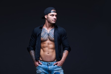 The man dressed blue jeans, black shirt and black baseball cap with tattoos on the dark background.