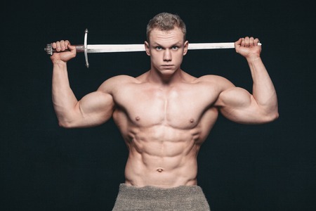 Bodybuilder man posing with a sword isolated on black background. Serious shirtless man...