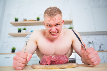 Muscular man with a naked torso in the kitchen with a piece of beef meat, the concept of a healthy diet. Athletic way of life. Predatory agressive degeneration of the face.の写真素材