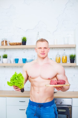 Muscular man with a naked torso in the kitchen with a salad and a piece of beef meat, the concept of a healthy diet. Athletic way of life.の写真素材