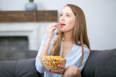 Beautiful young smiling woman watching a movie in the bed and eating popcorn.の写真素材